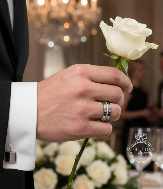 A hand wearing a wedding ring holds a white rose in front of a blurred background with a chandelier and floral arrangements.