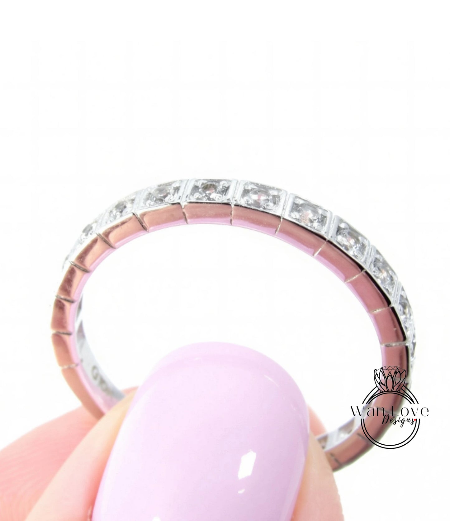 A close-up of a person's hand holding a pink nail polish bottle, with a ring featuring a row of diamonds on the finger.