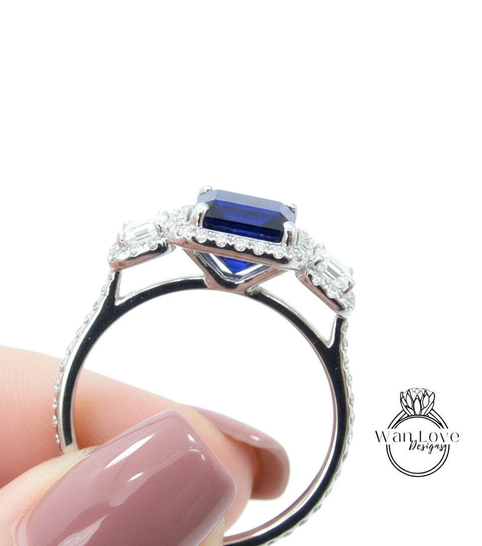 A close-up view of a person's hand holding a silver ring with a large blue gemstone, likely a sapphire, surrounded by smaller diamonds.
