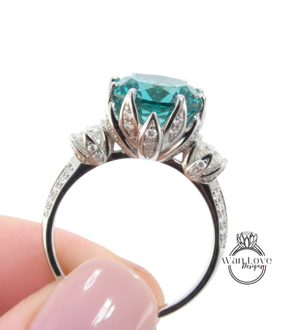 A close-up of a person's hand holding a silver ring with a large, turquoise-colored gemstone, likely a diamond, surrounded by smaller diamonds.