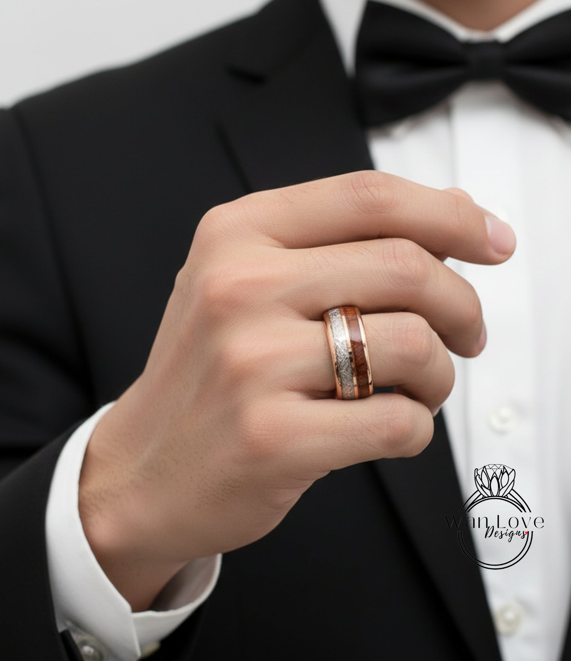 A close-up of a person's hand wearing a gold ring on the ring finger, with a black suit jacket and a black bow tie visible in the background.