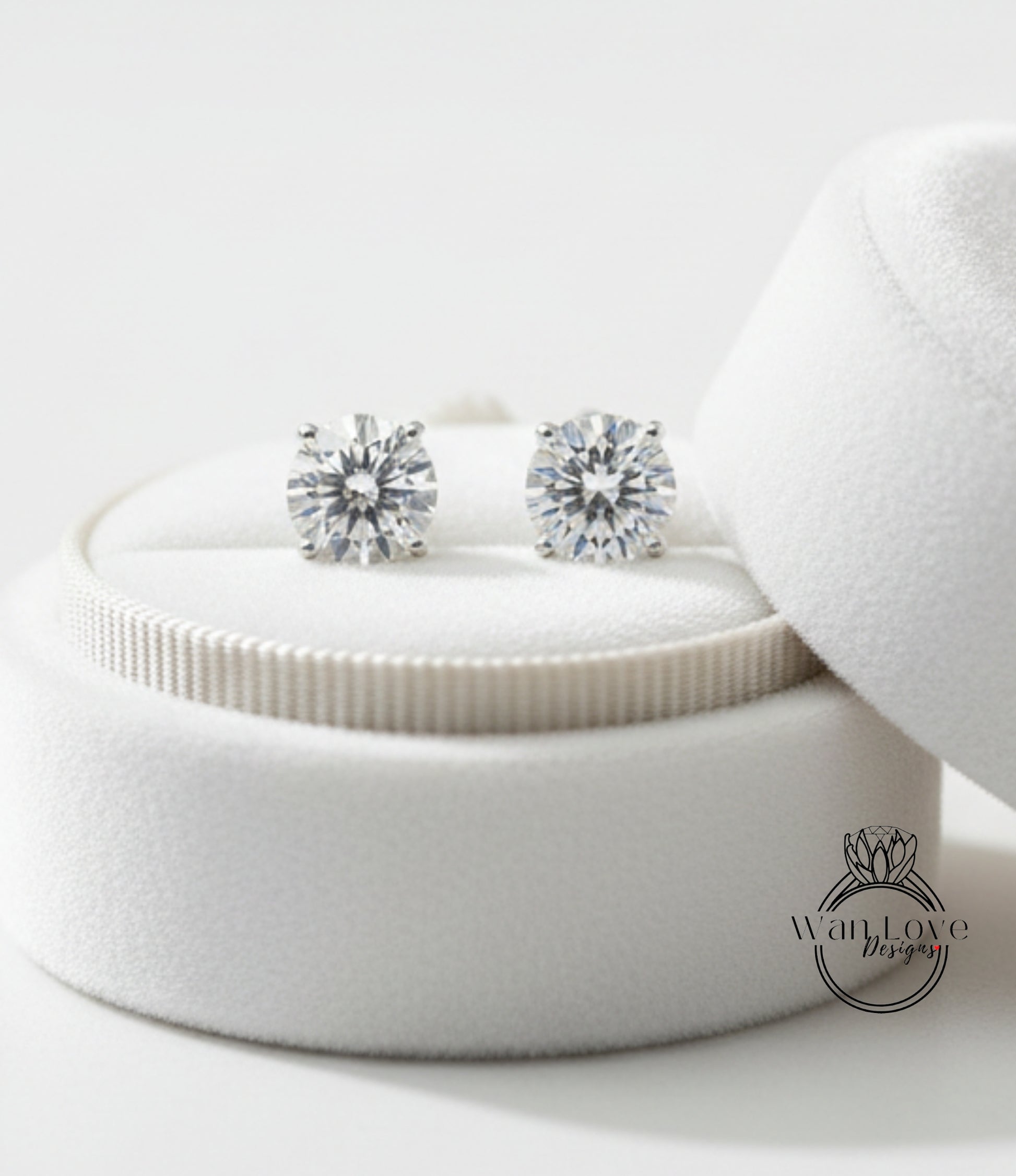 A pair of elegant diamond earrings displayed in a white velvet box, with a logo for "WanLove Designs" visible in the bottom right corner.