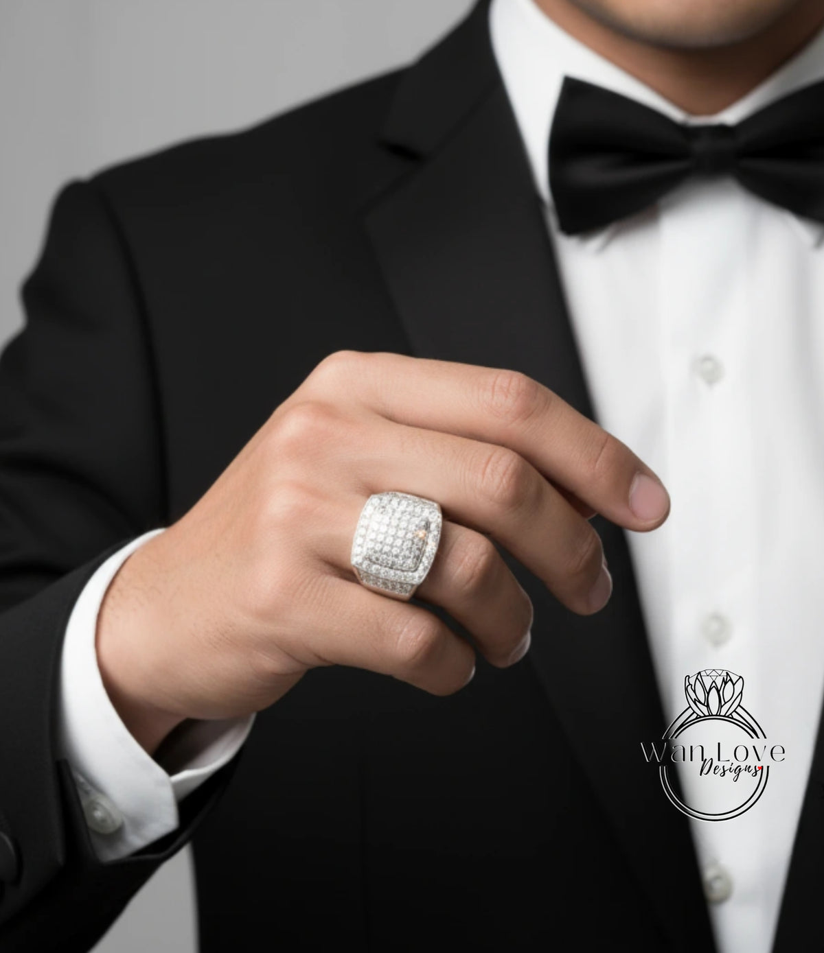 A close-up of a man's hand wearing a large diamond ring, with a black suit and bow tie visible.