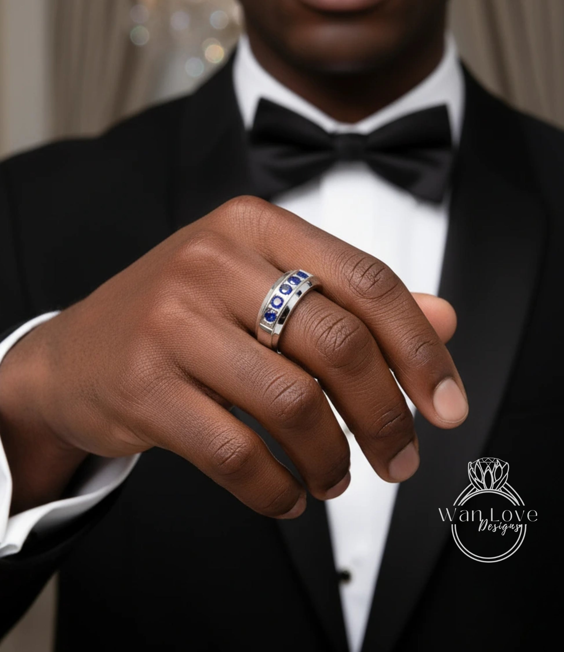 A close-up of a person's hand wearing a ring with blue gemstones, wearing a black tuxedo with a bow tie.