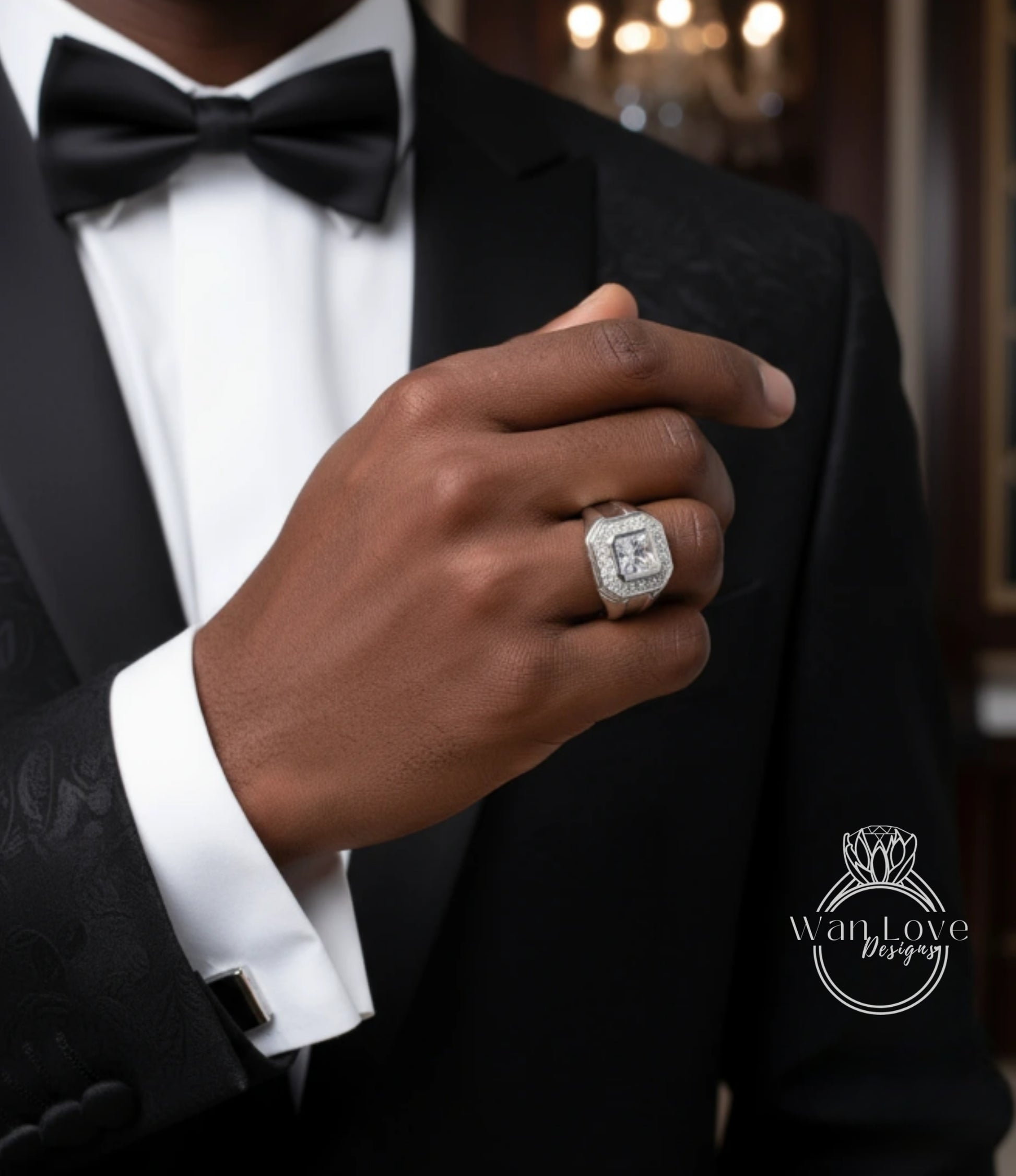 A close-up of a person's hand wearing a diamond ring, with a black bow tie and a black suit jacket.