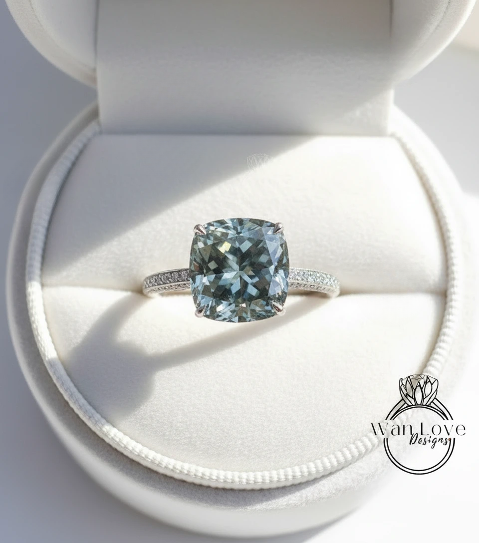 A large, blue-green gemstone engagement ring is displayed in a white jewelry box, showcasing its intricate design and sparkling details.