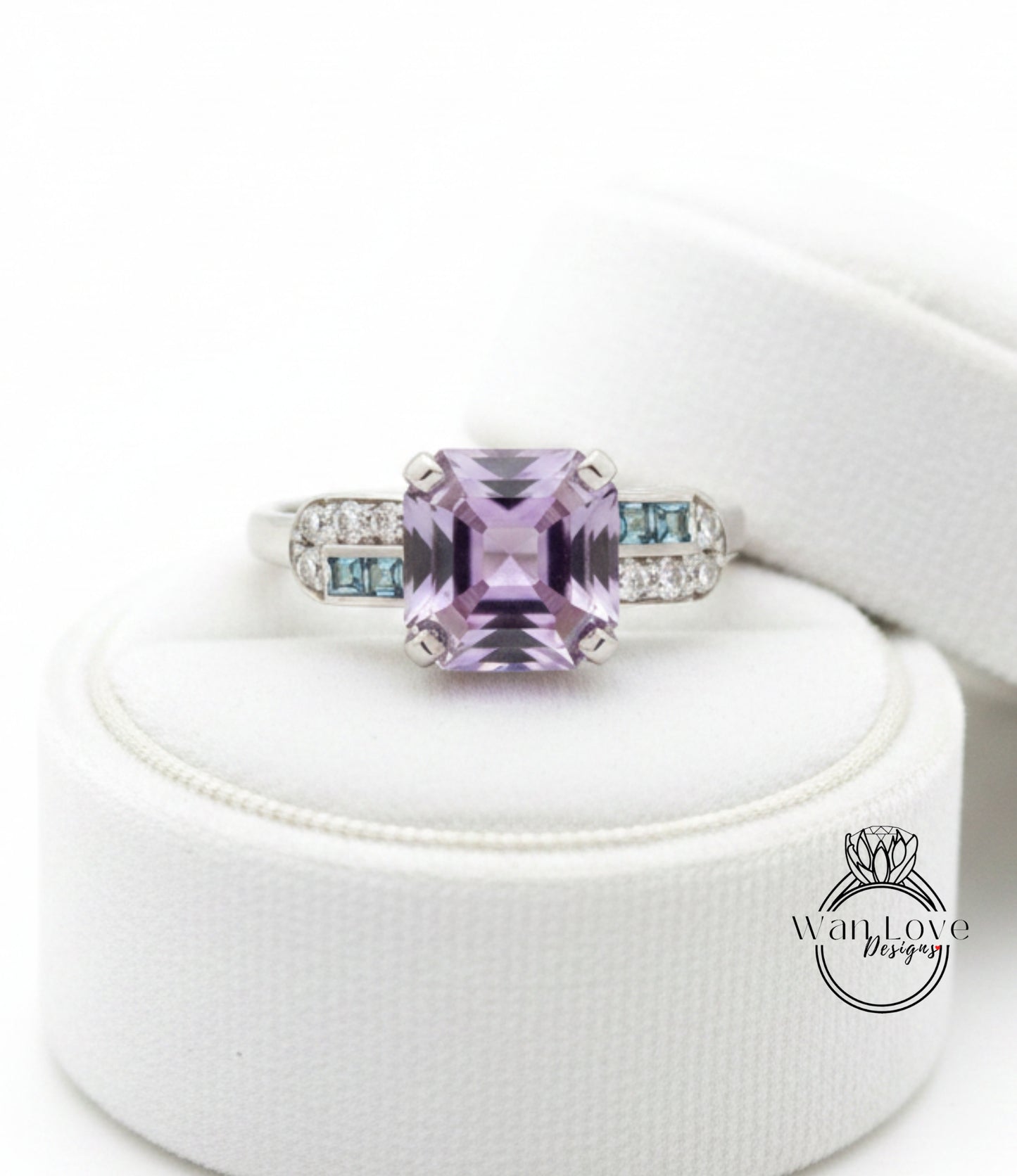 A ring with a large purple gemstone, possibly a purple sapphire, surrounded by smaller blue gemstones. The ring is displayed on a white stand, and there is a logo or text visible in the image.