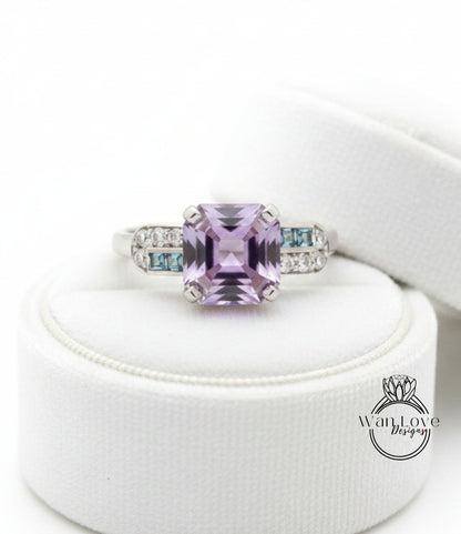 A ring with a large purple gemstone, possibly a purple sapphire, surrounded by smaller blue gemstones. The ring is displayed on a white stand, and there is a logo or text visible in the image.