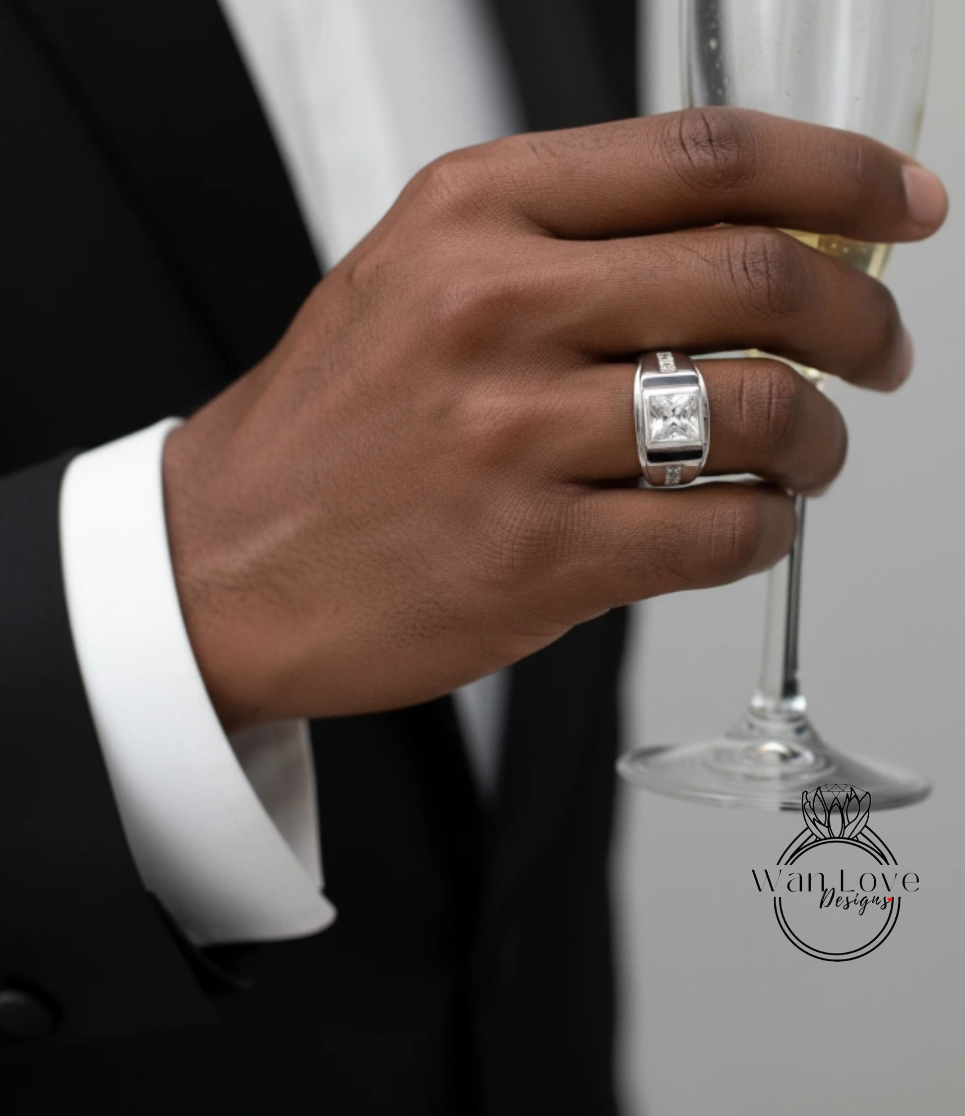 A close-up of a person's hand wearing a ring, holding a glass of champagne, and wearing a black suit jacket.