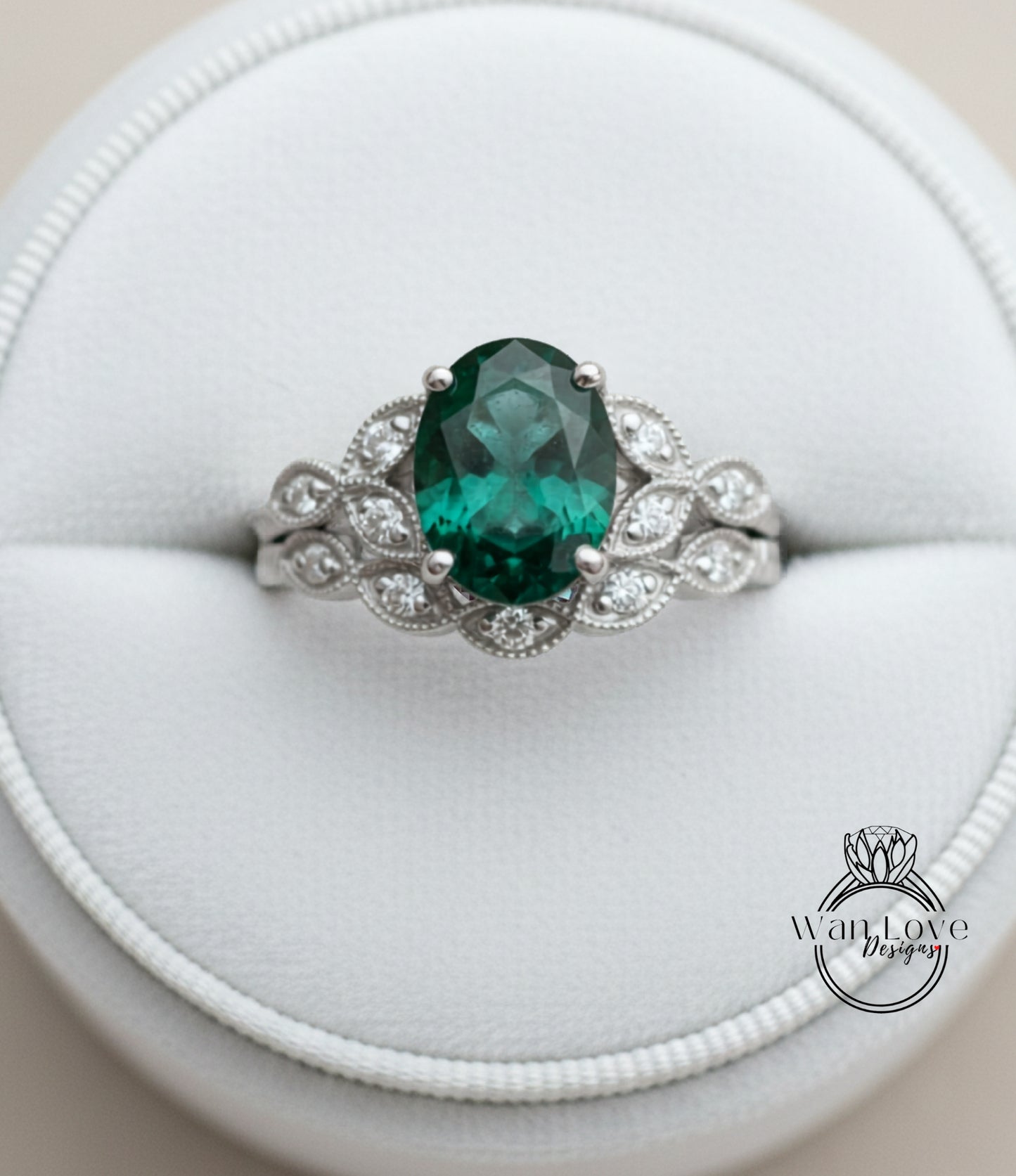 A silver ring with a large oval-shaped green gemstone, surrounded by smaller diamonds, placed in a white jewelry box.