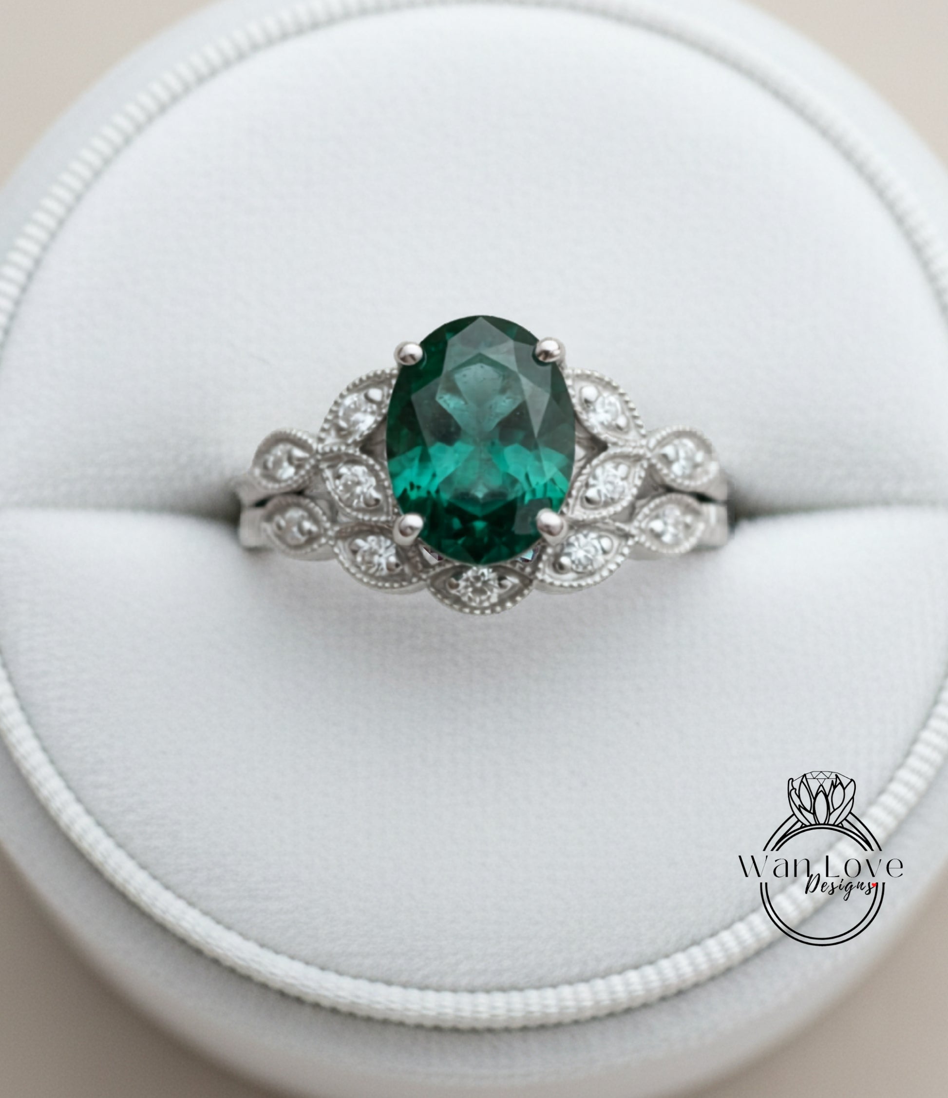 A silver ring with a large oval-shaped green gemstone, surrounded by smaller diamonds, placed in a white jewelry box.
