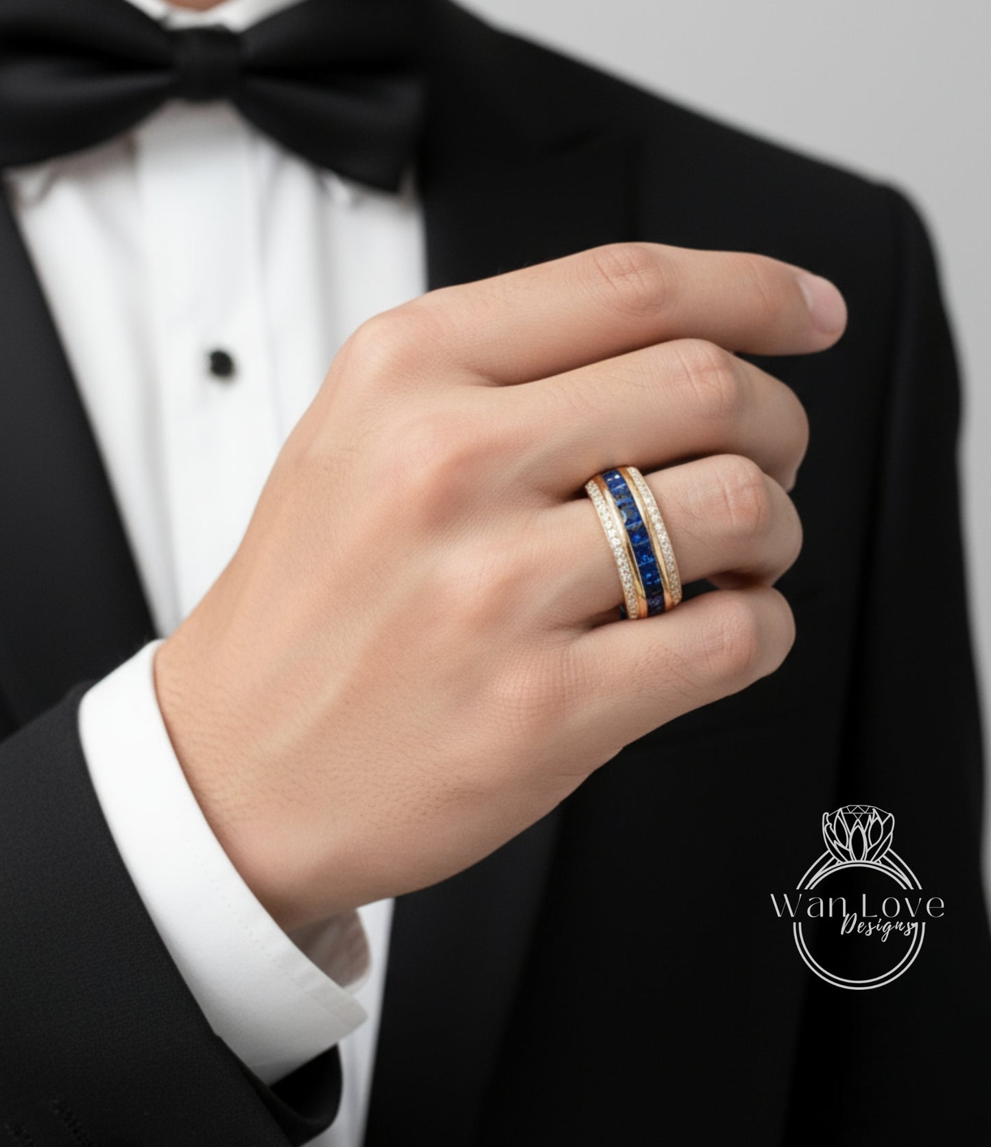 A close-up of a person's hand wearing a gold ring with blue gemstones, wearing a black tuxedo jacket.