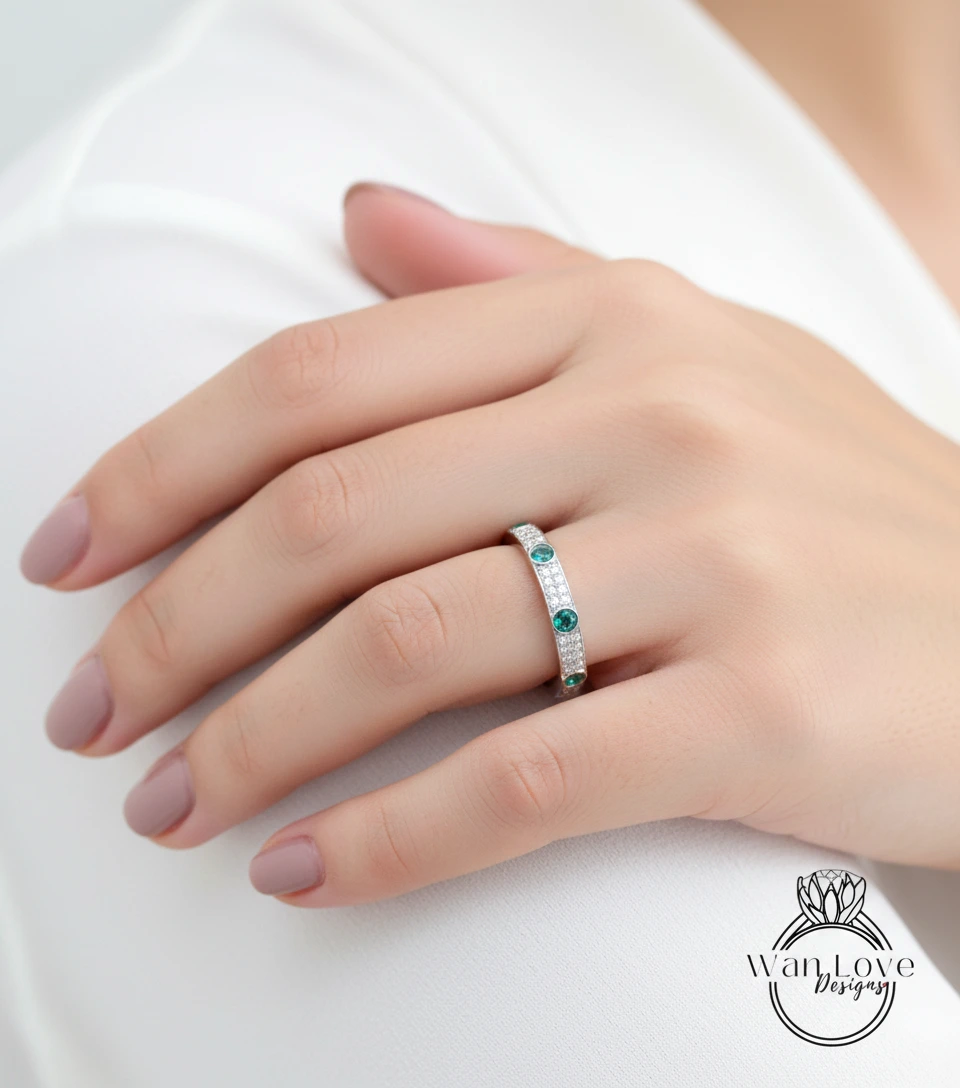 A close-up of a person's hand wearing a ring with a band of diamonds and green gemstones.