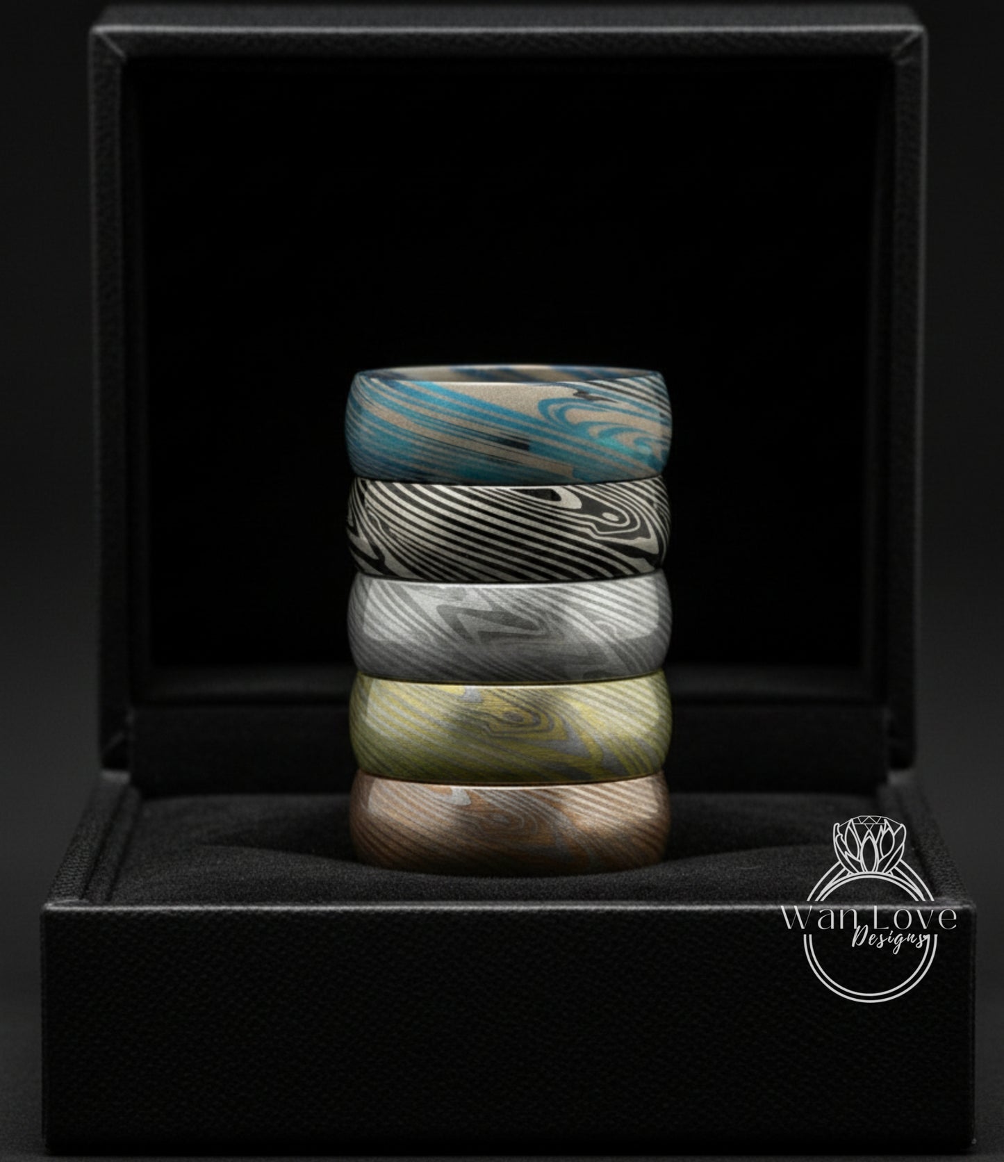 A stack of colorful rings is displayed in a black jewelry box against a dark background.