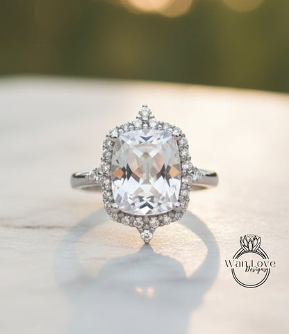 A large, clear gemstone ring with a halo of smaller diamonds around the center stone, set in a silver band, is displayed on a white surface.