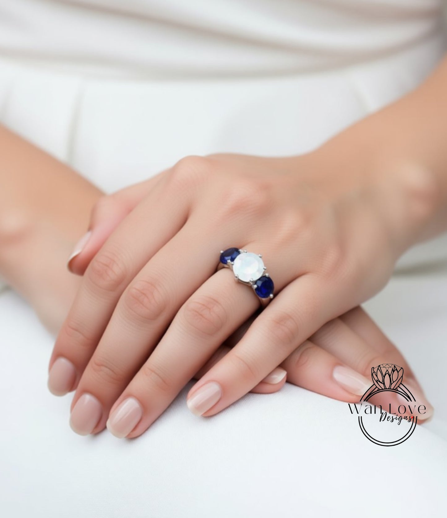 A close-up of a person's hand wearing a ring with a large white gemstone and two smaller blue gemstones.