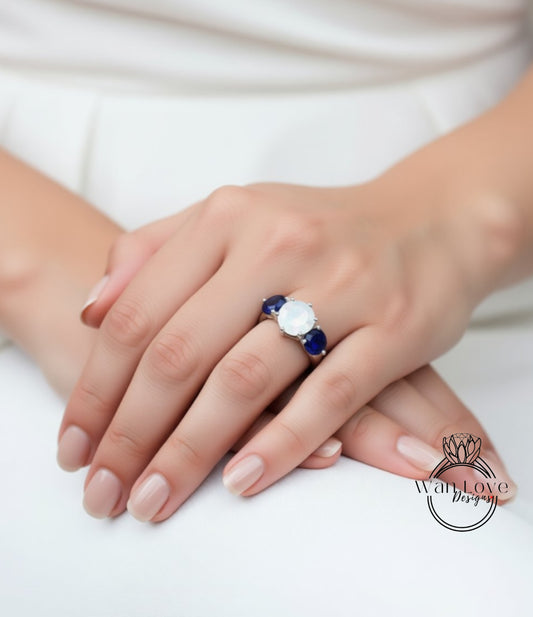 A close-up of a person's hand wearing a ring with a large white gemstone and two smaller blue gemstones.
