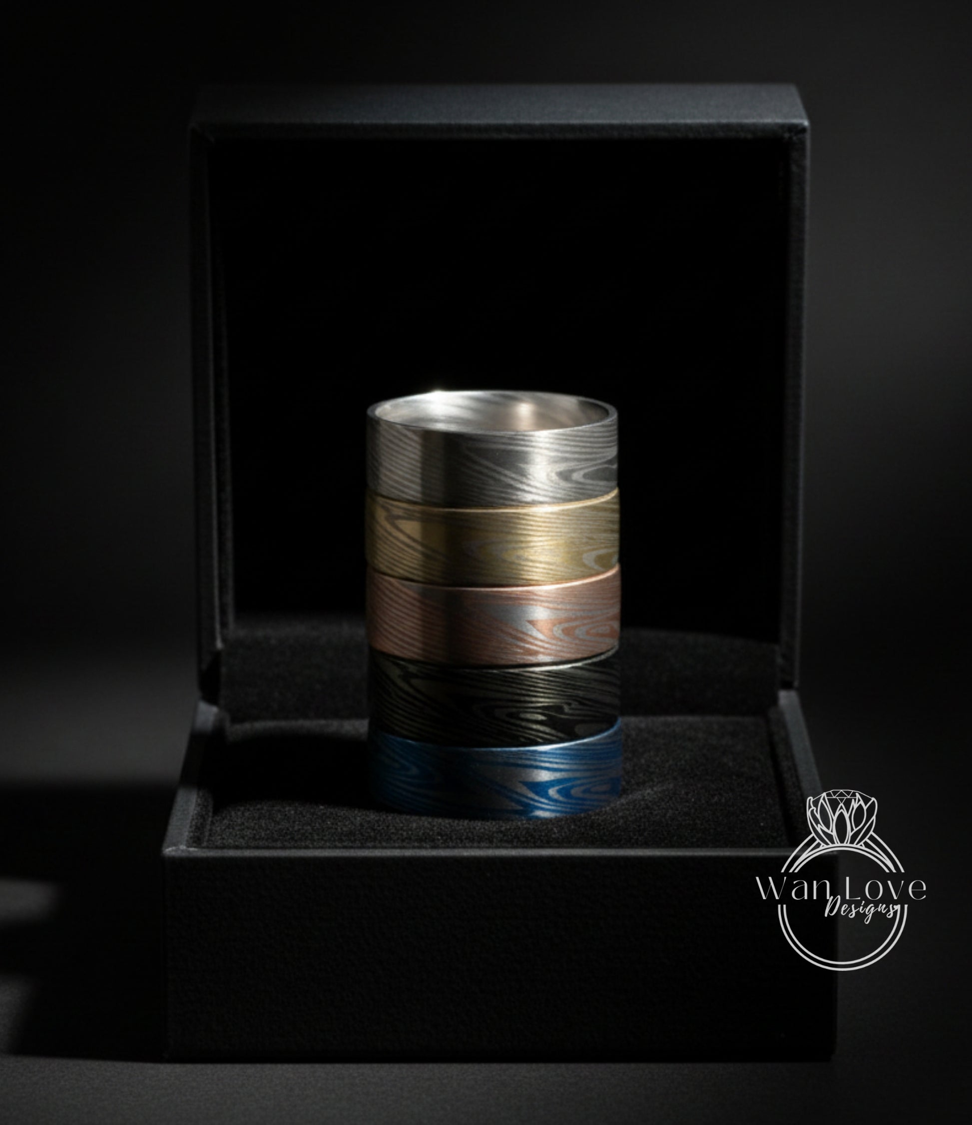 A black jewelry box containing a stack of rings in various colors, including silver, gold, blue, and brown.