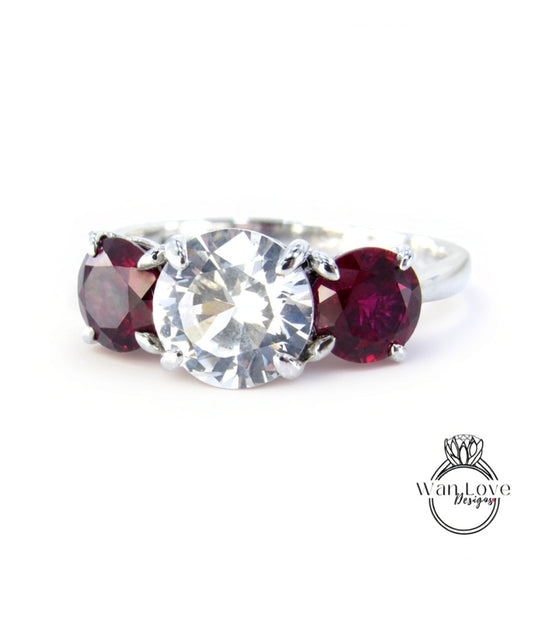A close-up image of a ring with a large, clear gemstone surrounded by two smaller, deep red gemstones.