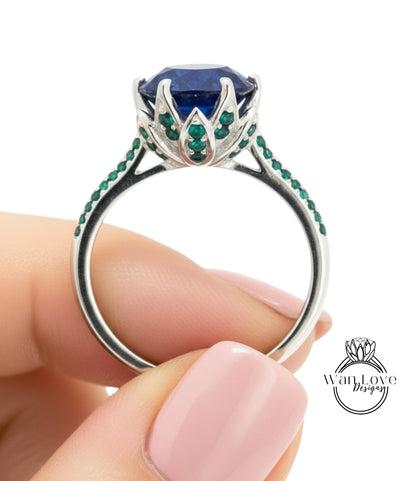 A hand holding a silver ring with a large blue gemstone and green gemstones.