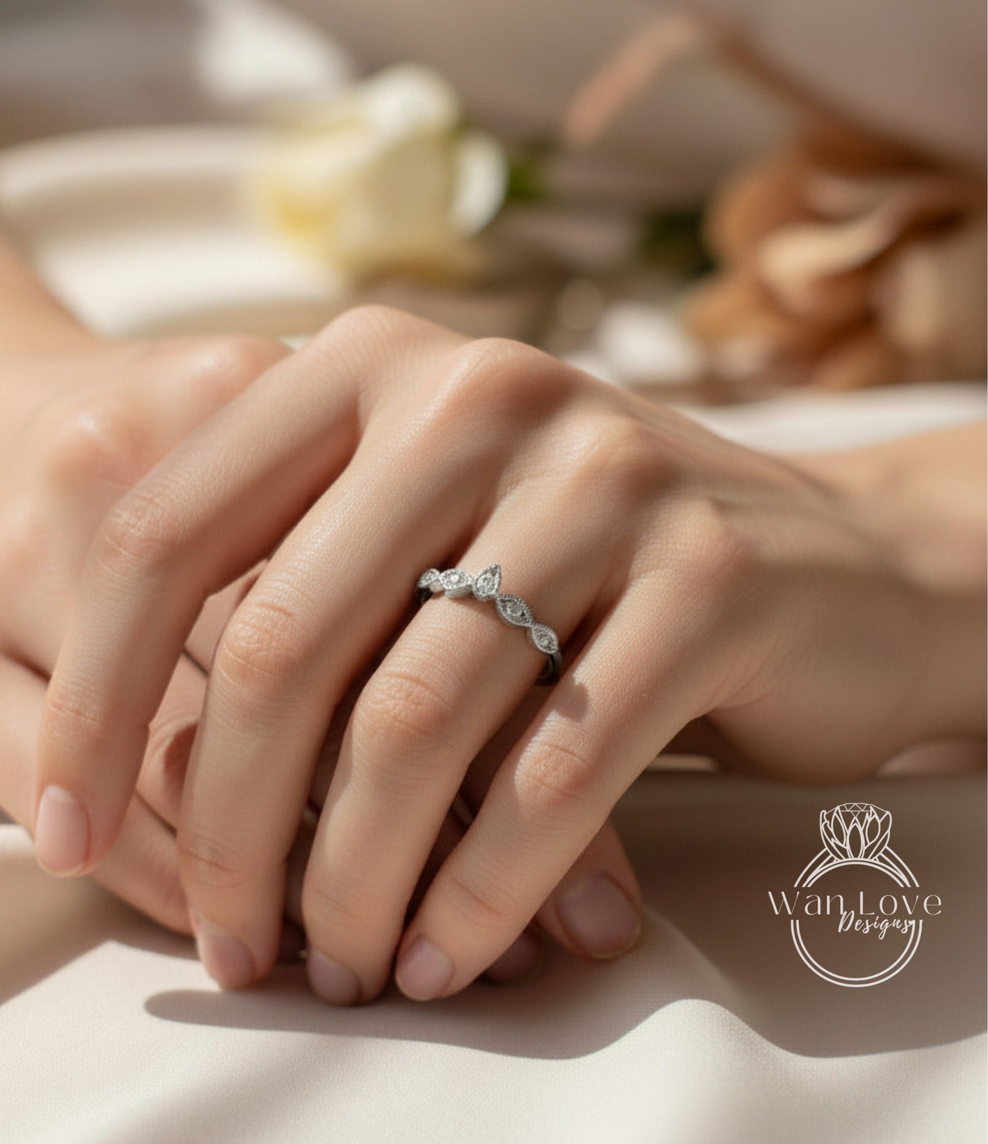 A close-up of a person's hand wearing a diamond ring, with a blurred background that includes a white rose and other floral elements.