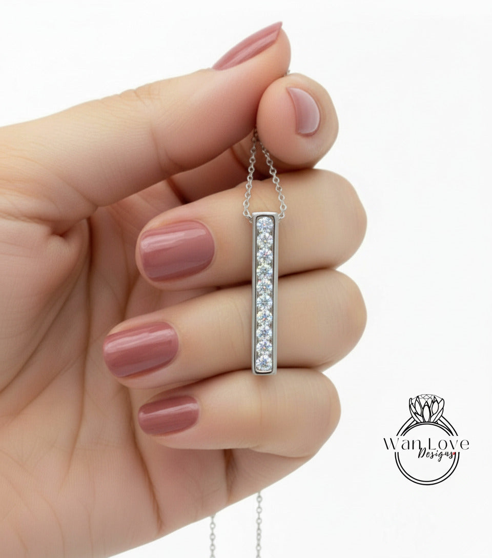 A hand holding a silver necklace with a vertical pendant, featuring a row of small diamonds.