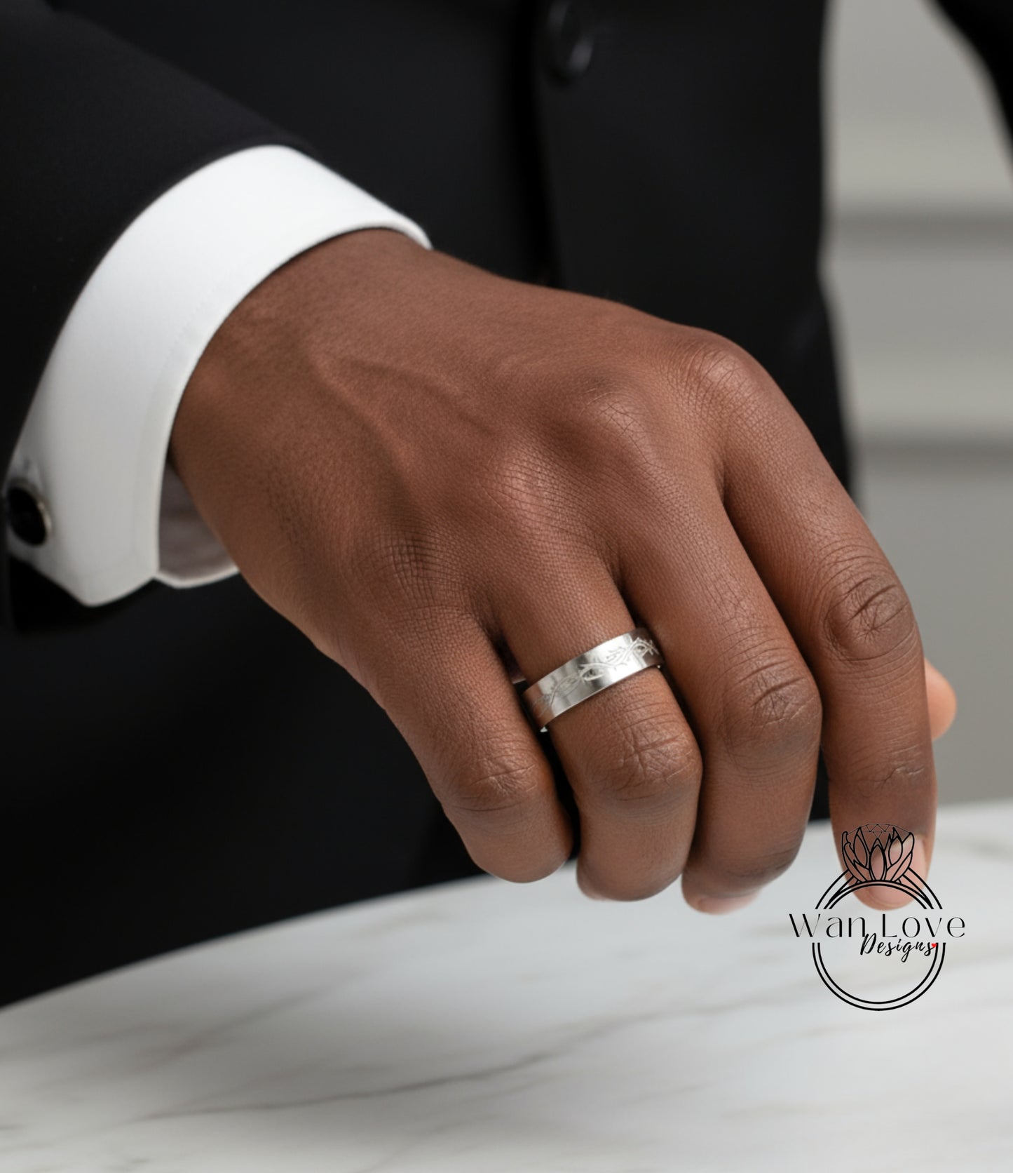 A close-up of a person's hand wearing a silver wedding ring, with a black suit jacket visible in the background.