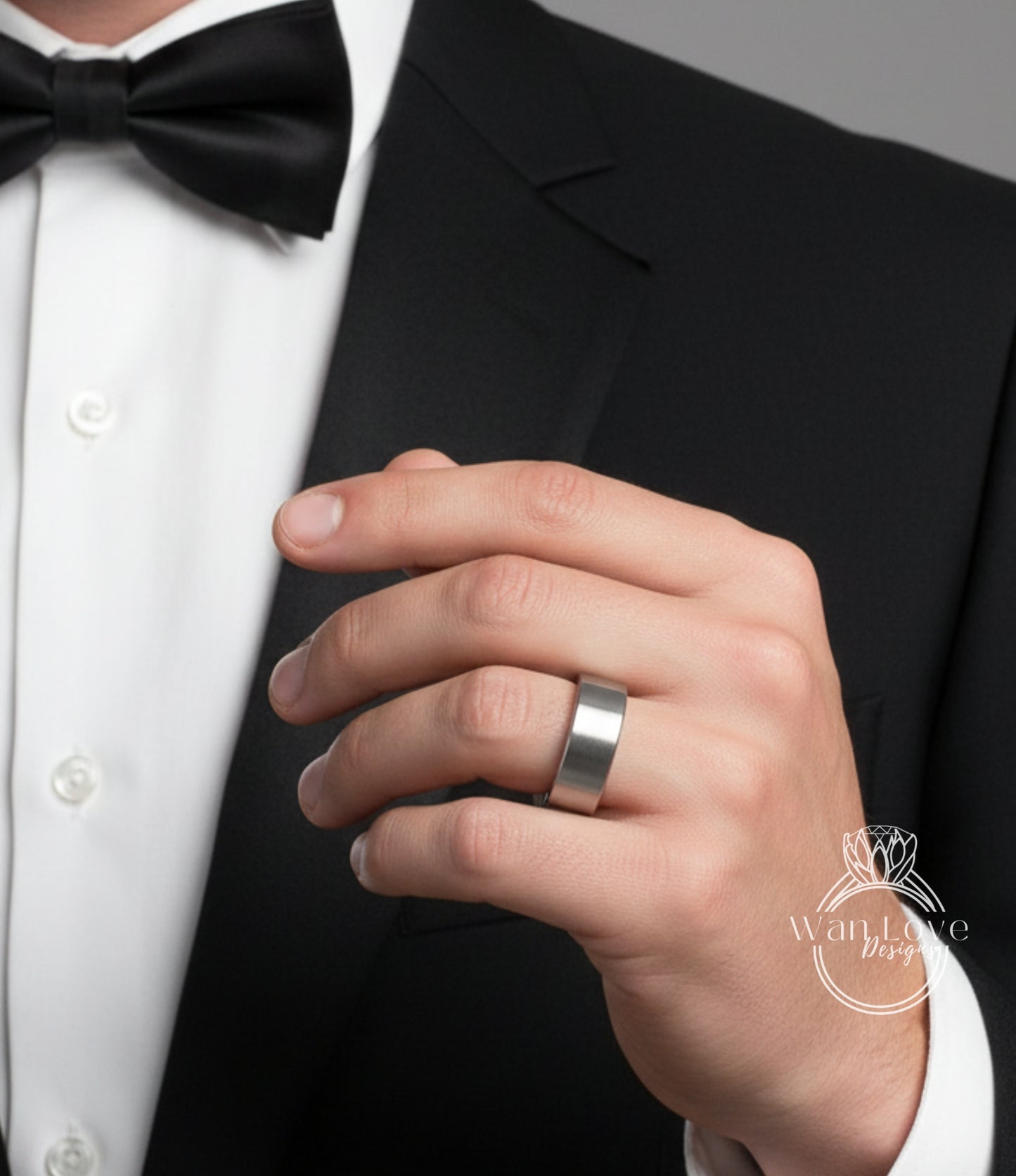 A close-up of a person's hand wearing a silver ring, with a black bow tie and a black suit jacket.