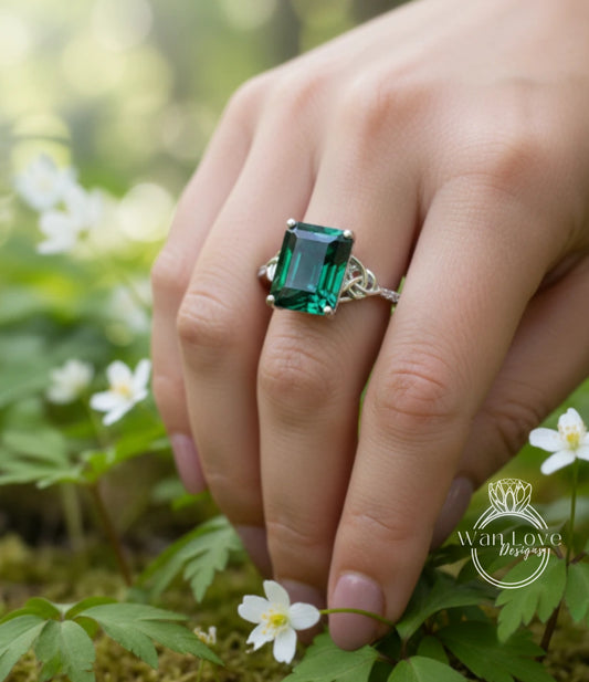 A close-up of a hand wearing a ring with a large, green gemstone, surrounded by white flowers and lush greenery.