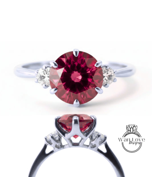 a stunning engagement ring with a large, deep red gemstone set in a silver band. The ring is adorned with three smaller diamonds, adding to its elegance and sophistication. The image showcases the ring from various angles, highlighting its intricate design and the beauty of the gemstone.