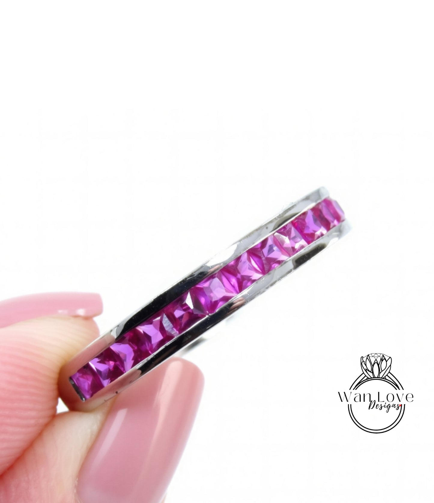 A close-up image of a person's hand holding a ring with a row of pink gemstones, likely rubies, set in a silver band.