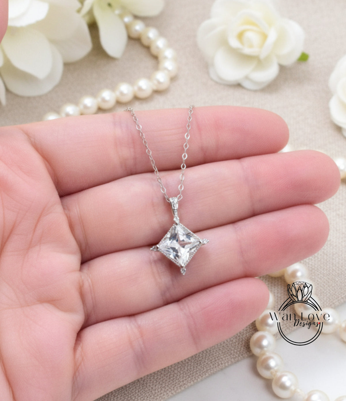 A hand holding a delicate necklace with a clear gemstone pendant, surrounded by white pearls and a floral arrangement.