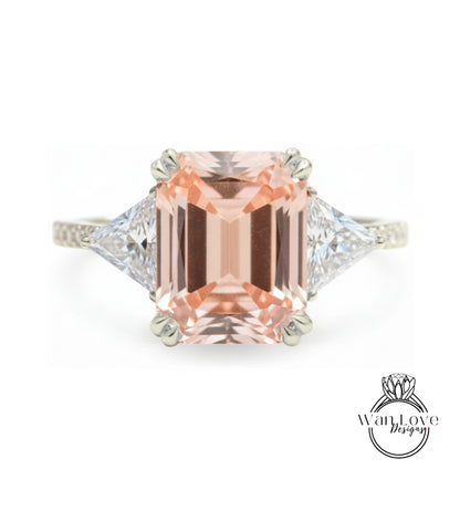 A large, peach-colored gemstone engagement ring with a diamond-encrusted band and two triangular-shaped diamonds on either side.