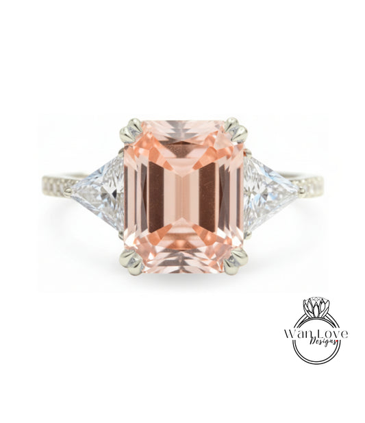 A large, peach-colored gemstone engagement ring with a diamond-encrusted band and two triangular-shaped diamonds on either side.