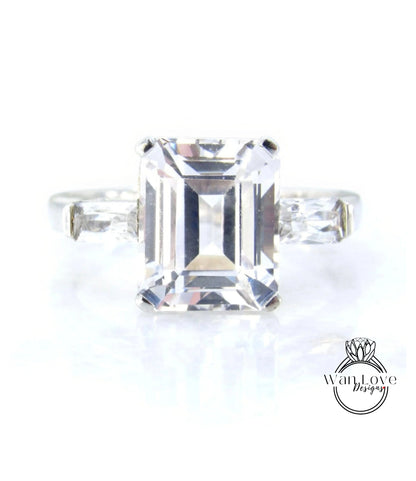A large, clear gemstone ring with a diamond-like center stone surrounded by smaller diamonds on either side.
