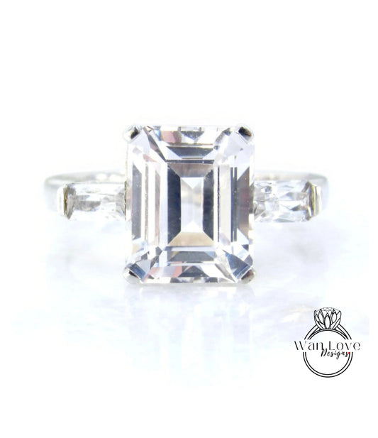 A large, clear gemstone ring with a diamond-like center stone surrounded by smaller diamonds on either side.