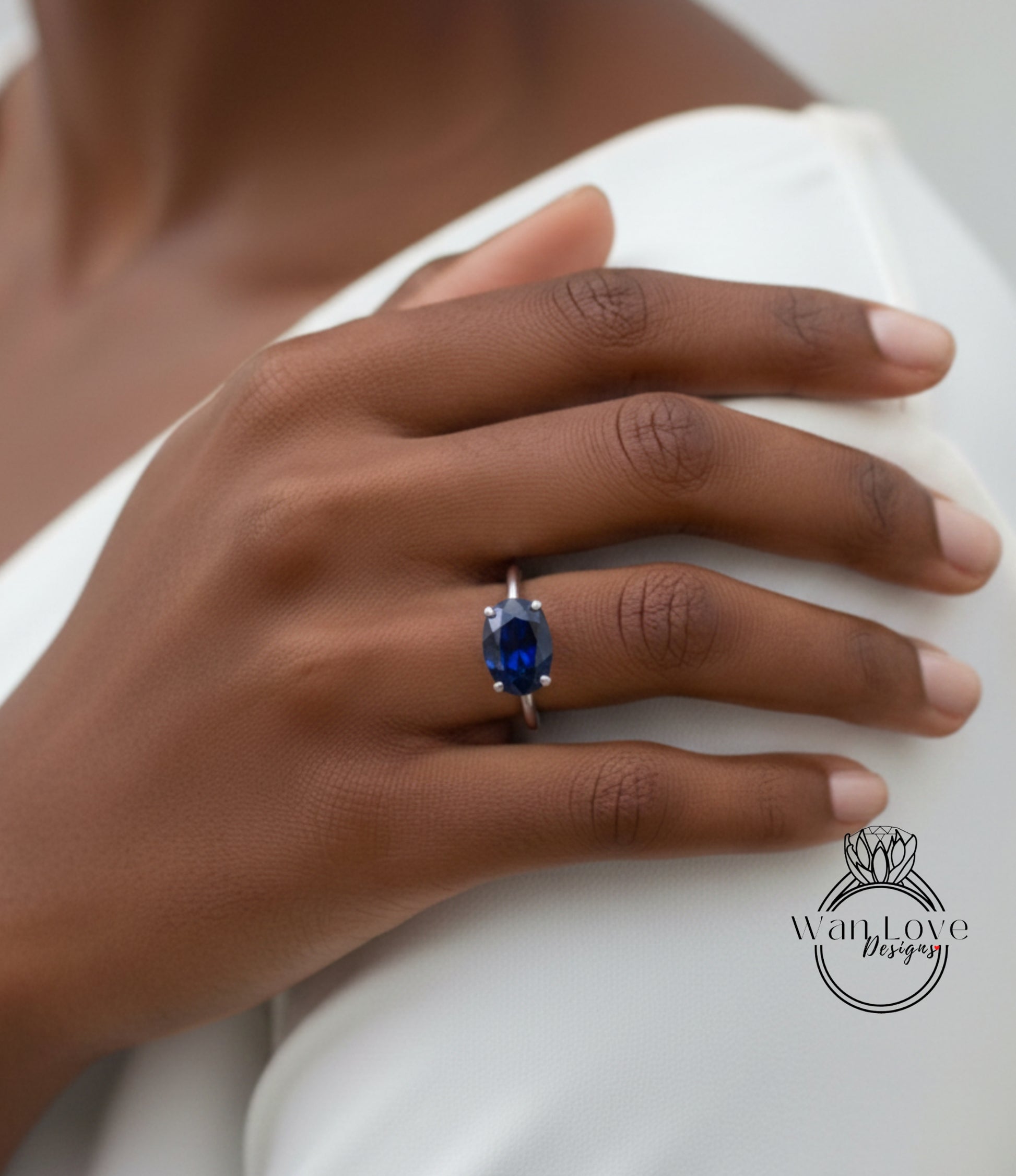 A close-up of a hand wearing a ring with a blue gemstone, resting on a white surface.