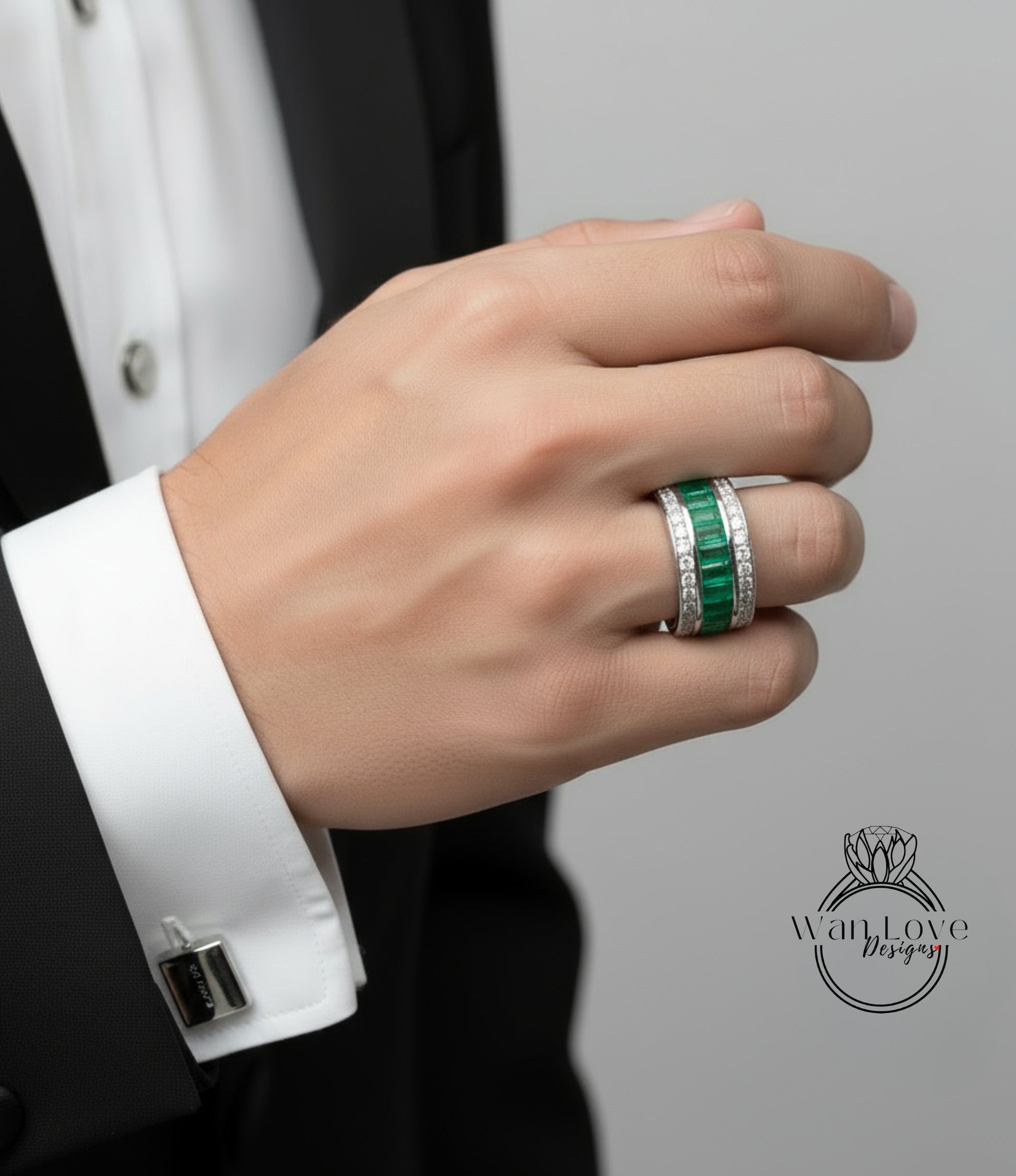 A close-up of a person's hand wearing a ring with green gemstones, a cufflink, and a black suit jacket.