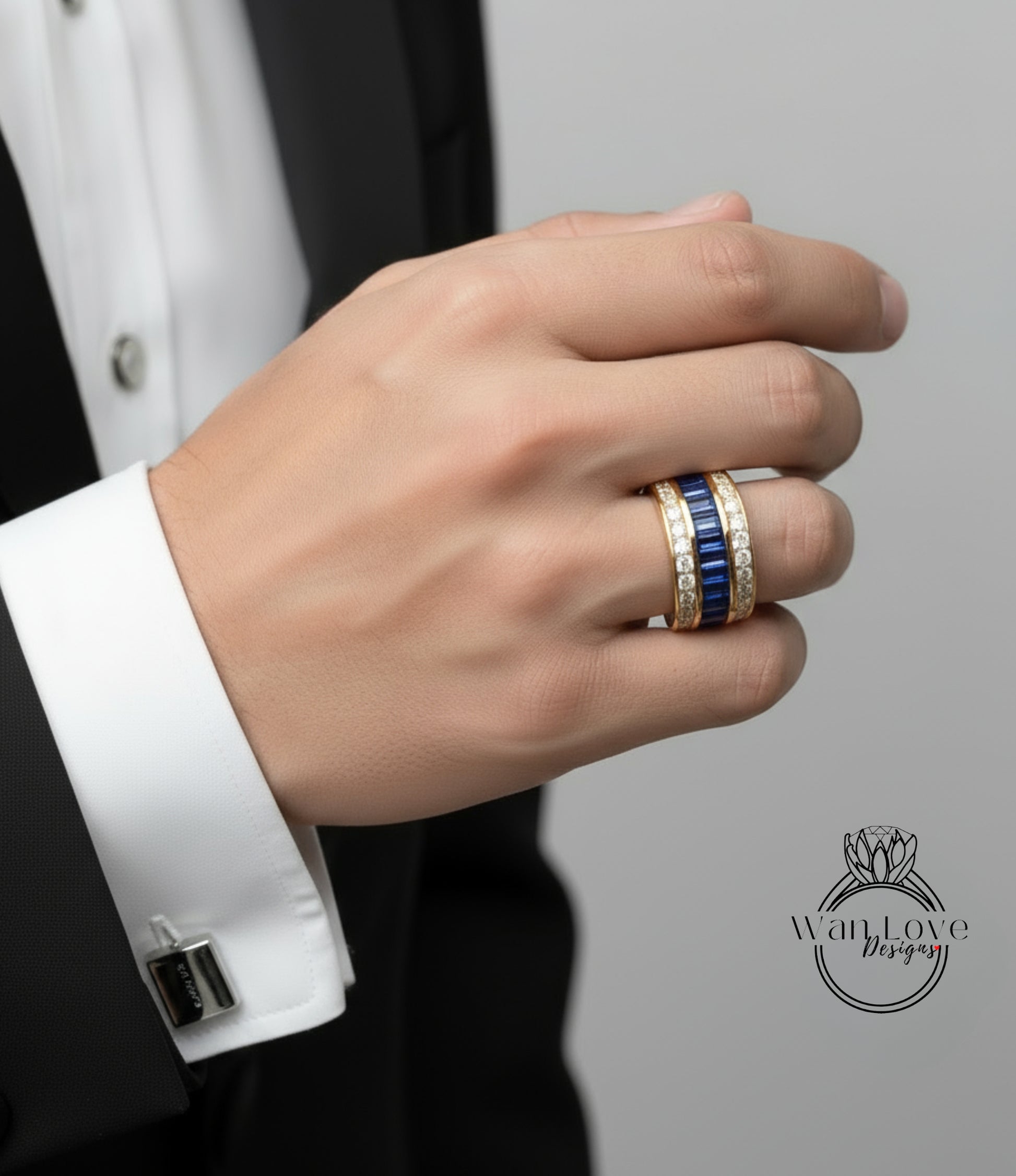 A close-up of a person's hand wearing a gold ring with blue gemstones, wearing a black suit jacket, and a white shirt cuff.