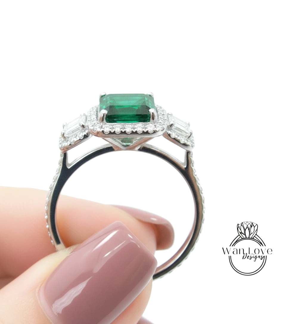 A close-up of a person's hand holding a silver ring with a large, square-cut emerald gemstone set in a halo of smaller diamonds.