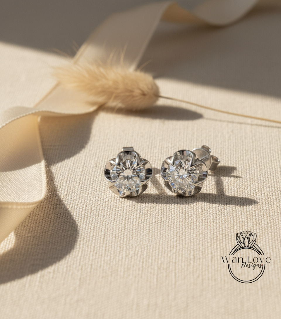 A pair of diamond earrings with a large central diamond and smaller diamonds surrounding it, placed on a beige fabric surface with a ribbon and a small flower in the background.