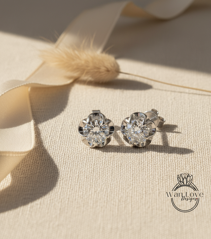 A pair of diamond earrings with a large central diamond and smaller diamonds surrounding it, placed on a beige fabric surface with a ribbon and a small flower in the background.
