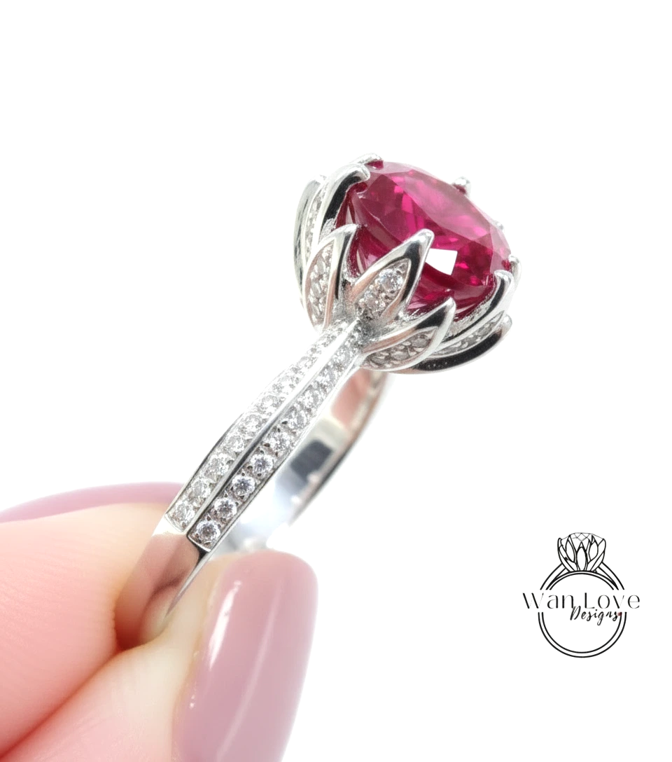 A close-up of a person's hand holding a silver ring with a large, vibrant red gemstone, likely a sapphire or ruby, surrounded by smaller diamonds.