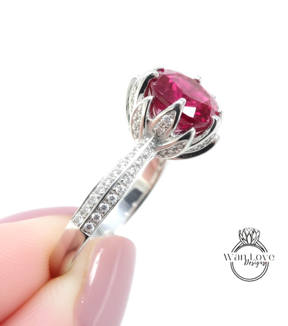 A close-up of a person's hand holding a silver ring with a large, vibrant red gemstone, likely a sapphire or ruby, surrounded by smaller diamonds.