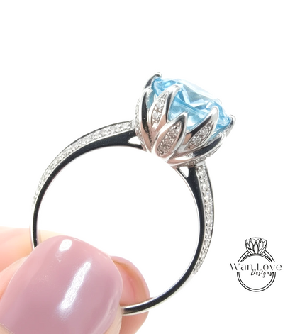 A close-up image of a person's hand holding a silver ring with a large blue gemstone, likely an aquamarine or blue topaz stone.